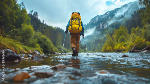 Solo hiker with a backpack using trekking poles to cross a mountain river, surrounded by a misty forest landscape.