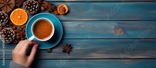 A warm and inviting autumn or winter scene with a cup of coffee cinnamon sticks sugar orange slices and pine cones arranged on a wooden background Blue mittened hands hold the coffee cup creating a c