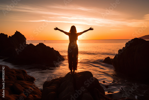 Silhouette of a beautiful woman standing on a rock with her arms open facing the sea at sunset