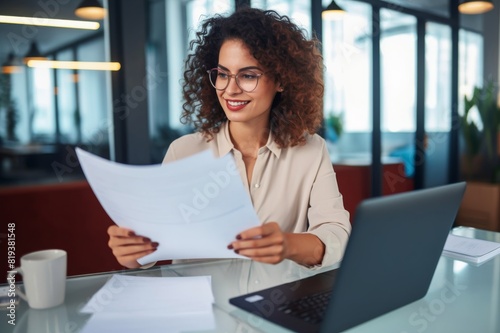 Young happy and successful businesswoman in glasses working with documents inside office, Hispanic woman with laptop looking at bills and contracts, financier with curly hair using laptop Young happy 