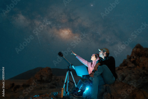 heterosexual couple sitting in the desert watching the stars and Milky Way next to a telescope, stargazing and exploration concept 