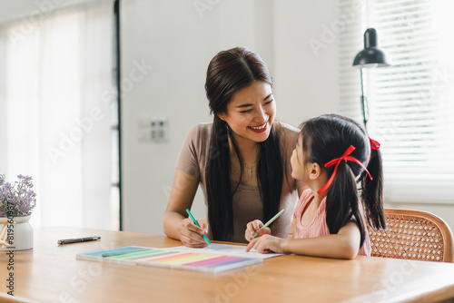 Young mother assists her daughter with coloring activities at a home desk during daytime.