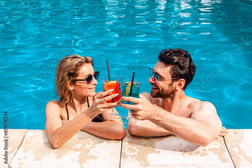 Happy couple in trendy sunglasses with refreshing beverages sitting in a holiday resort swimming pool. Leisure and summer vacation concept