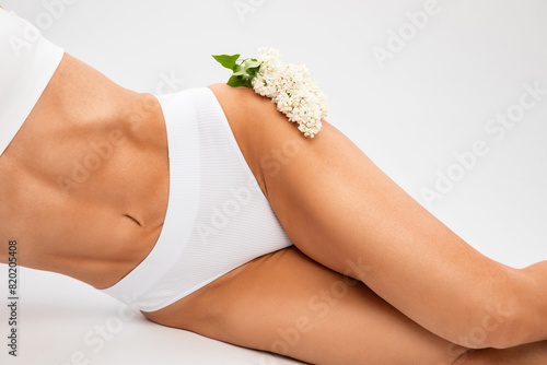 A slender swarthy woman holding a white branch of lilac flowers in a white underwear on a white background. Healthy lifestyle, sport and diet.