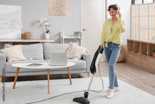 Beautiful young woman in headphones cleaning carpet with handheld vacuum cleaner in living room