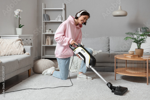 Happy young woman in headphones cleaning carpet with handheld vacuum cleaner in living room