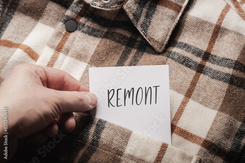 White card with a handwritten inscription "Remont", held in the hand against the background of a brown plaid shirt (selective focus), translation: renovation