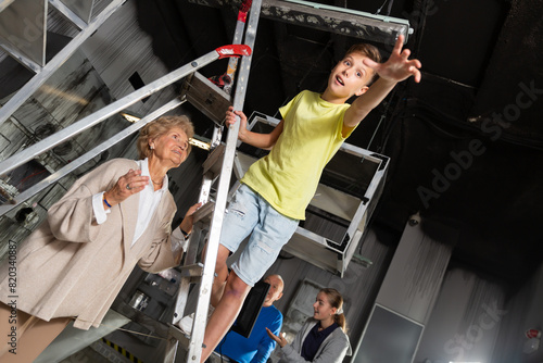 Young boy standing on stepladder and reaching his hand in escape room. His grandmother standing next to him and holding stepladder.