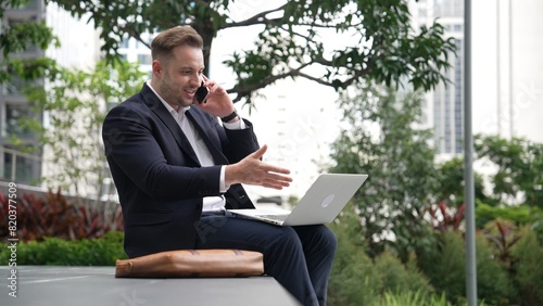 Caucasian businessman sitting at eco park while talking by using phone and working on laptop. Professional executive manager explain marketing idea from computer while calling to investor. Urbane