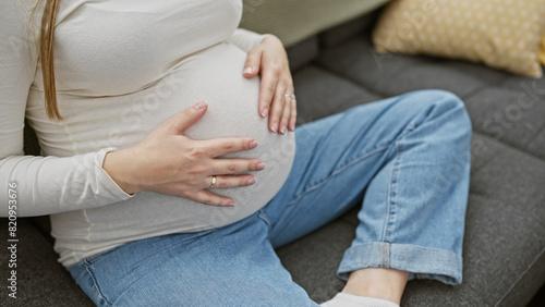 A pregnant hispanic woman in casual wear, resting on a couch indoors with a hand on her belly, embodying motherhood.