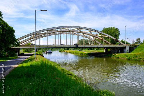 Photo of a metal curved bridge over the canal (Netekanaal in Viersel, Belgium). Blue sky with clouds.