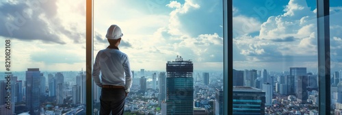 A male engineer in a hard hat stands contemplating the cityscape from a high vantage point in an office