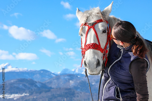 A girl in sunglasses with a white horse wearing a red bridle against a background of mountains and a blue sky