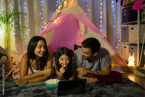 Cheerful family watching a movie at home with popcorn, surrounded by cozy lights and a fun teepee backdrop