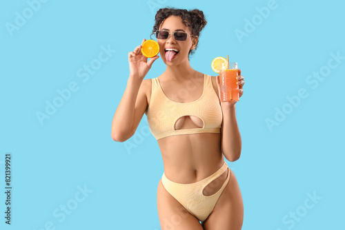 Young African-American woman in swimsuit with cocktail and citrus showing tongue on blue background