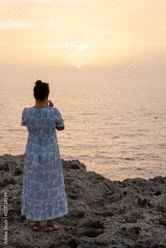 Rear view of a woman in her 30s wearing a white and blue maxi dress, capturing a sunset over the sea on a rocky shore with her phone, in Menorca, Spain