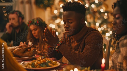 A religious African male prays before eating a turkey dinner with friends and family at a festive table with family. A young black man thanks God, thanks Jesus Christ, before he eats food during the