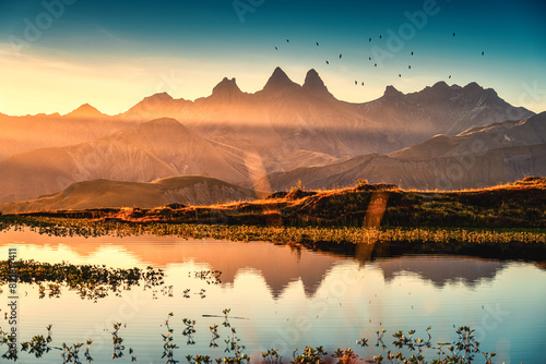 Sunrise shines over Lac Guichard with bird flying on Arves massif and lake reflection at Aiguilles d'Arves, French Alps, Savoie, France