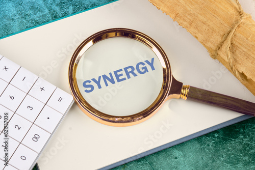 Synergy, cooperation or efficiency in business concept. The word synergy through a magnifying glass on a blank sheet of notepad next to a calculator and a roll of papyrus