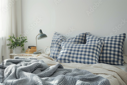 A bed with blue and white checkered pillows and a grey blanket on top is viewed from the front, with a lamp placed next to a bedside table which has books.