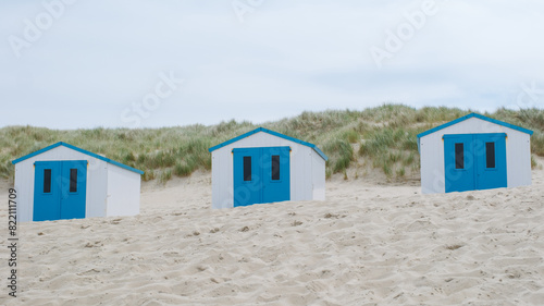 Three charming beach huts with distinctive blue doors stand out on the sandy shore of Texel, Netherlands.