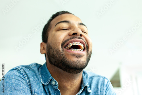 African American man with braces smiles and covers his smile and laughs, man laughs with his eyes closed, close-up