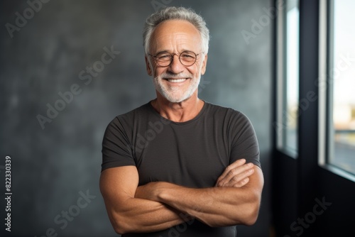 Portrait of a smiling man in his 60s with arms crossed while standing against bare monochromatic room