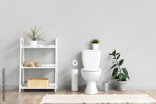 Interior of restroom with toilet bowl, shelving unit and plant near grey wall