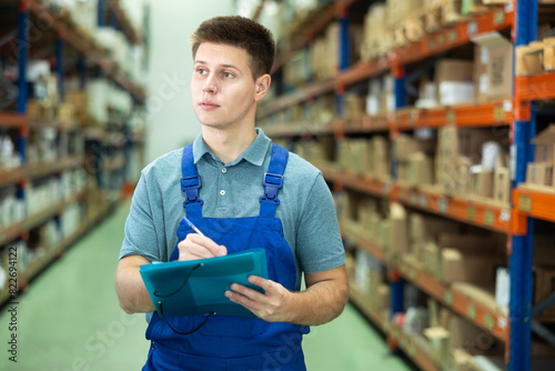 In warehouse rack area of paint store, guy worker in blue overall checks availability and quantity of goods and receipt documents for products