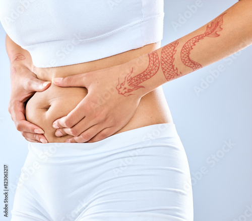 Woman, hands and belly with holding stomach for diet results isolated on studio background. Tattoo, fitness and abdomen with stubborn fat, cellulite and tummy tuck in sportswear for training