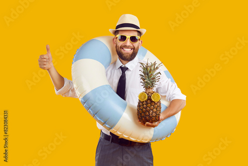 Portrait of happy funny man in sunglasses and beach hat wearing office clothes with rubber ring and fresh pineapple isolated on studio yellow background showing thymb up. Summer holiday trip concept.