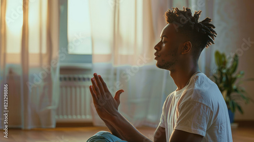 Focused calm African American guy meditating at home, practicing pranayama, breath work, keeping Namaste greeting hands gesture, sitting on floor with closed eyes. Side view.