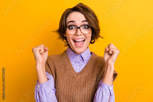 Portrait of ecstatic woman wear knit waistcoat in glasses clenching fists win betting scream yeah isolated on yellow color background