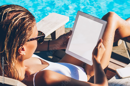 Female freelancer in sunglasses sunbathing on sunbed near blue pool and doing distance job online on modern tablet connected to 4G internet during summer trip thanks to hot tours and freelance work