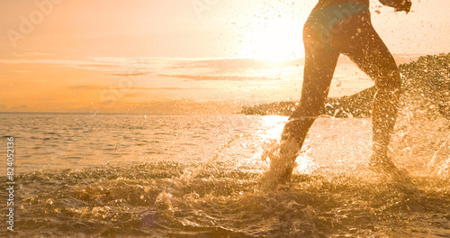 LENS FLARE, CLOSE UP, SILHOUETTE: young woman splashes water while running through shallow seawater along beautiful beach in golden sunset light. Dynamic and fun sports activities on summer holidays.