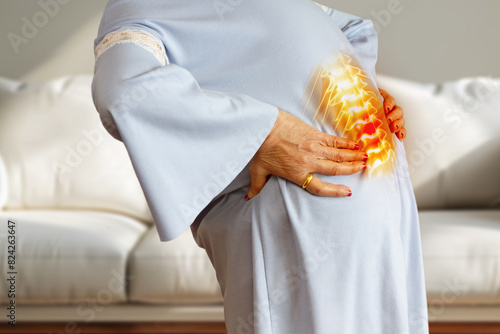 Elderly woman wrapping her waist with her hands in the living room of her house. Spinal disk disorder, back pain concept. 