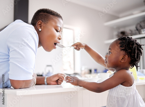 Black child, feeding and mother with spoon in kitchen for food, meal or share together at home. Young African, little girl or kid giving mom a taste of cereal bowl for breakfast, fiber or calcium