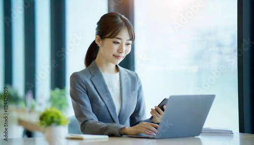 都会のオフィスの窓辺で、スーツを着た若い女性が、スマートフォンとパソコンを使って仕事をしている。