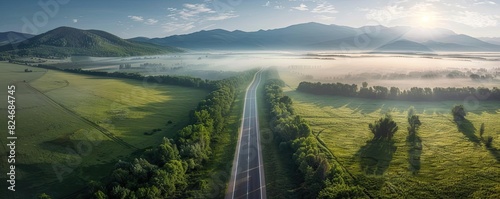 Aerial view of a straight country road through lush green fields and mountains in the background on a misty morning.