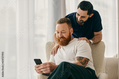 Two men in casual clothes sit in a chair, bonding as they look at a cell phone together in a modern living room.