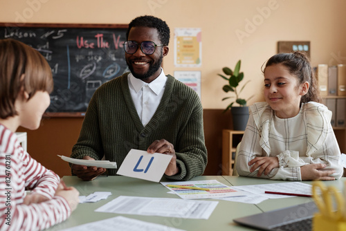 Cheerful African American male teacher of English working with group of kids showing phonics cards during lesson in classroom