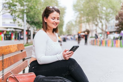 A woman sits to rest on a bench in the street and looks at her phone smiling