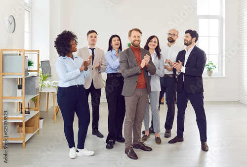 Group of company employees congratulating their happy male colleague on job well done applauding standing in office. Business people and group of staff clapping to young smiling man.