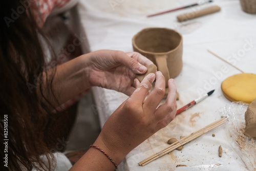 A close-up of the hands of a young woman as she shapes a heart out of clay to decorate a mug. Master class on pottery in a creative workshop. Valentines day creative hobbies.