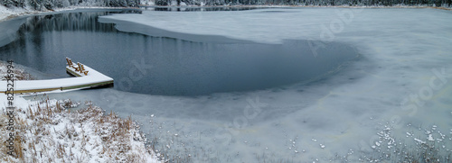 Panoramic view of a small lake with a fishing dock as the water in the lake is freezing into ice. An area of open water still exists in front of the dock. 