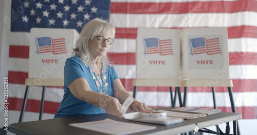 Three quarter angle, medium shot, elderly woman election worker checks over forms and stickers for voters at polling station. US flag and voting booths behind