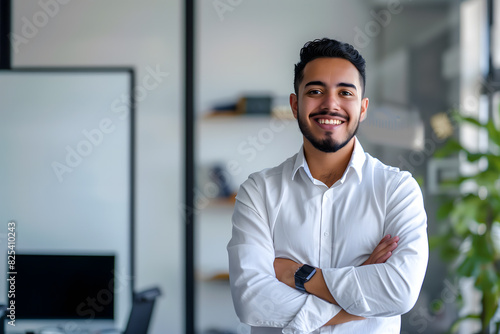 smiling young Hispanic business man standing with arms crossed in an office, whiteboard in the background
