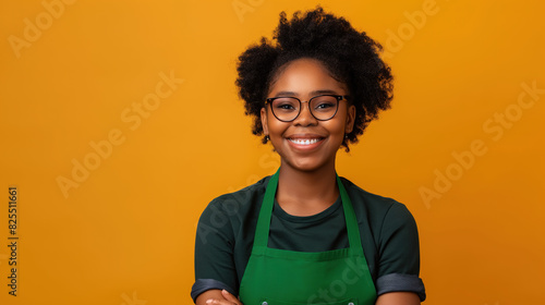 happy african american young woman wearing green apron and glasses, looking at camera isolated on orange background with copy space for your design