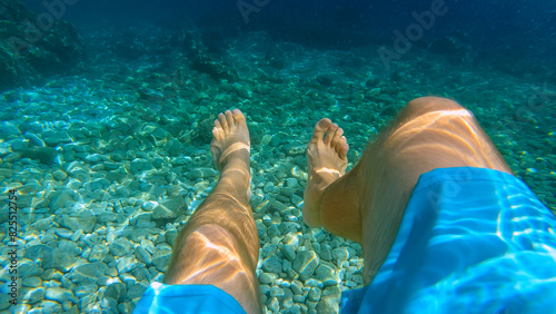POV UNDERWATER: Pair of male legs floating over a pebble seabed in a crystal sea. Young man in swimming shorts enjoys a refreshing swim in astonishingly blue Adriatic Sea during his summer holidays.