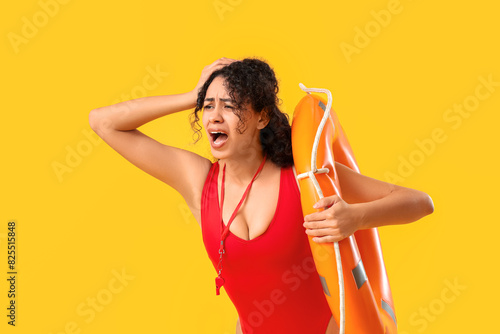 Beautiful young shocked African-American female lifeguard with ring buoy screaming on yellow background